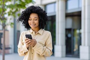 jovem africano americano mulher com telefone sorridente, empresária lado de fora escritório construção usa Smartphone aplicativo, tipos mensagens e navega Internet Páginas. foto