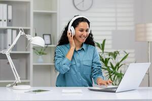 feliz mulher goza ouvindo para música enquanto trabalhando em dela computador portátil dentro uma bem iluminado casa escritório ambiente, exibindo produtividade e conforto. foto