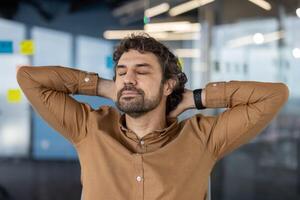 uma meia idade homem com encaracolado cabelo relaxa de alongamento dele braços atrás dele cabeça dentro uma contemporâneo escritório atmosfera, demonstrando uma momento do estresse alívio. foto