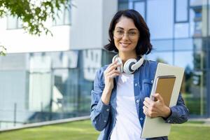 uma confiante jovem mulher vestindo óculos e fones de ouvido detém uma caderno, em pé lado de fora uma moderno universidade prédio, simbolizando Educação e tecnologia. foto