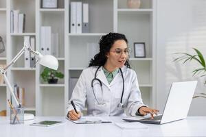 sorridente latina médico dentro uma laboratório casaco acionado dentro dela trabalhos às a computador dentro uma moderno clínica escritório. foto