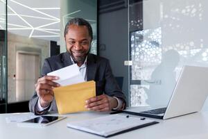 africano americano homem de negocios lendo Boa notícias, homem dentro escritório local de trabalho segurando enviar envelope carta com Boa notícias, patrão dentro o negócio terno maduro e adulto. foto