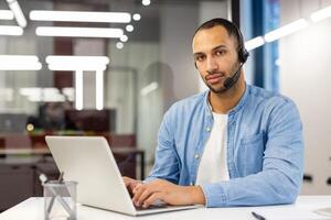 retrato do uma sério hispânico homem dentro uma camisa sentado dentro a escritório, às a mesa dentro frente do a computador portátil e com confiança olhando às a Câmera.. foto