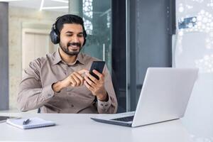 satisfeito bem sucedido hindu homem às local de trabalho dentro escritório, homem com telefone dentro mãos e fones de ouvido assistindo conectados e ouvindo para música audio podcasts, trabalhador usando computador portátil às trabalhar. foto