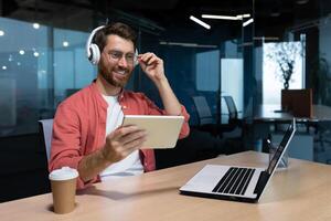 bem sucedido homem de negocios às local de trabalho sorridente homem assistindo conectados sentado às escrivaninha vestindo fones de ouvido e óculos programador segurando tábua computador dentro escritório. foto