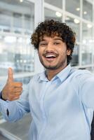 uma alegre jovem homem com encaracolado cabelo dá uma polegares acima dentro uma moderno escritório contexto, exalando confiança e positividade. perfeito para o negócio e motivacional temas. foto