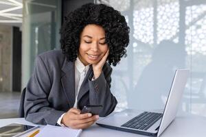 bem sucedido empresária às local de trabalho dentro escritório usando telefone, fêmea trabalhador lendo mensagem e sorridente, africano americano mulher segurando Smartphone usando conectados aplicativo. foto