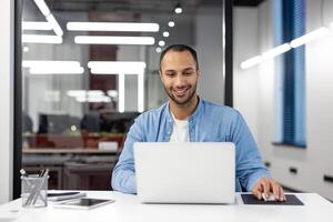 sorridente jovem profissional homem usando computador portátil às dele escrivaninha dentro uma bem iluminado moderno escritório, mostrando produtividade e conforto. foto