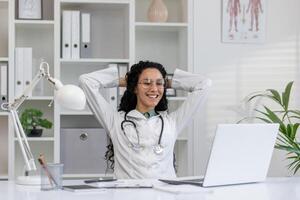 alegre latina médico dentro branco casaco levando uma pausa às dela escritório escrivaninha com computador portátil, sentindo-me conteúdo e feliz. foto
