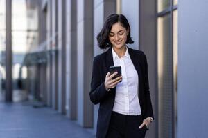 uma jovem lindo o negócio mulher é caminhando lado de fora a escritório prédio, uma mulher dentro o negócio roupas é sorridente, usando a inscrição em a telefone, navegando a Internet, digitando uma mensagem. foto