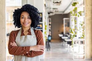 retrato do uma jovem sorridente afro-americano mulher quem trabalho dentro uma refeições estabelecimento, restaurante. em pé dentro a avental dentro de casa, olhando às a Câmera com cruzado braços. foto