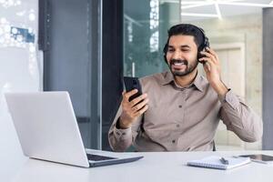 satisfeito bem sucedido hindu homem às local de trabalho dentro escritório, homem com telefone dentro mãos e fones de ouvido assistindo conectados e ouvindo para música audio podcasts, trabalhador usando computador portátil às trabalhar. foto