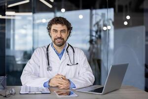 profissional masculino médico com uma estetoscópio por aí dele pescoço sentado dentro uma moderno clínica escritório, aparecendo confiante e acessível. foto