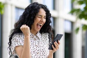 jovem alegre mulher vencedora recebido conectados notificação em telefone, hispânico mulher com encaracolado cabelo a comemorar sucesso e triunfo caminhando dentro cidade perto escritório construção fora. foto