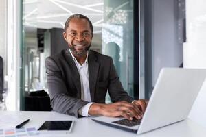 retrato do bem sucedido feliz africano americano chefe, homem sorridente e olhando às Câmera, homem de negocios dentro o negócio terno sentado às escrivaninha com computador portátil dentro escritório. foto