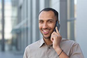 fechar-se foto do uma sorridente homem lado de fora a escritório construção alegremente falando em a telefone, uma homem de negocios dentro uma casual camisa hispânico olhando às a Câmera retrato