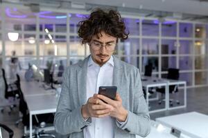 sério masculino empreendedor com encaracolado cabelo absorvido dentro lendo uma texto mensagem em dele telefone às uma corporativo escritório. foto