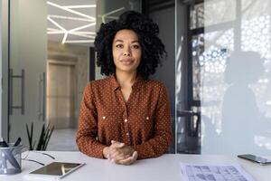 uma confiante jovem africano americano mulher dentro uma à moda polca ponto camisa poses às dela área de trabalho com uma tábua e projeto ,em uma bem iluminado moderno escritório contexto. foto