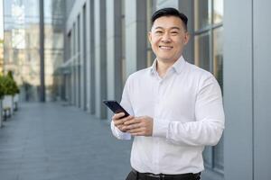 retrato do uma sorridente jovem ásia homem de negocios dentro uma branco camisa em pé fora, segurando uma Móvel telefone e olhando às a Câmera. foto