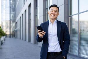 retrato do uma jovem ásia masculino homem de negocios em pé sorridente em a rua perto a escritório Centro, segurando uma telefone e olhando às a Câmera. foto