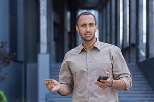 retrato do insatisfeito e enganado homem, homem de negocios lado de fora escritório construção olhando às Câmera e segurando banco crédito cartão e Smartphone foto