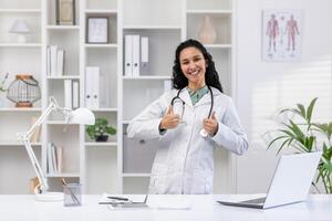 retrato do alegre bem sucedido sorridente fêmea médico dentro escritório às ambiente de trabalho, mulher dentro branco médico casaco em pé dentro escritório perto escrivaninha com computador portátil, mostrando polegares acima. foto