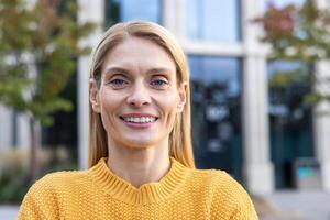 uma retrato do uma confiante meia idade empresária sorridente ao ar livre. ela é vestindo uma amarelo suéter, com uma moderno escritório construção dentro a fundo. foto