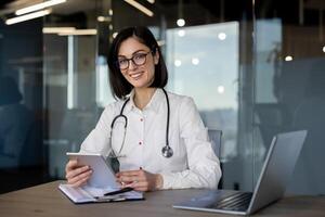 sorridente profissional mulher dentro médico casaco e estetoscópio segurando uma tábua, com computador portátil às ambiente de trabalho. foto