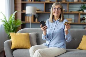 moderno consumismo conceito com uma mulher segurando crédito cartão e Smartphone para conectados compras enquanto sentado confortavelmente em uma sofá às lar. foto