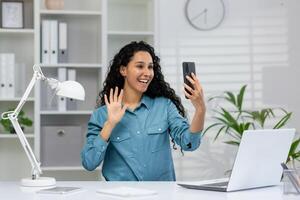 uma alegre mulher às uma moderno escritório escrivaninha é acenando Olá para alguém em uma ligar em dela Smartphone, aparentemente desfrutando uma casual conversação. foto