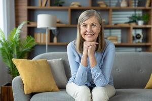 uma alegre Senior mulher com cinzento cabelo sorridente às a Câmera, sentado em uma sofá dentro uma bem decorado, moderno vivo quarto preenchidas com natural claro. foto