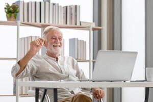 alegre Senior homem com computador portátil e café copo em mesa desfrutando dentro vivo quarto foto