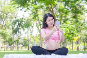 ásia grávida mulher usando uma Smartphone falando em uma chamar, jovem ásia mulher relaxante dentro a parque foto
