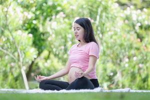 ásia grávida mulher meditando enquanto sentado dentro uma lótus posição dentro a parque, meditando em maternidade foto
