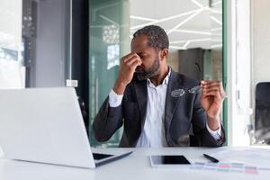 sobrecarregado homem às local de trabalho dentro escritório, africano americano homem de negocios trabalhando sentado com computador portátil às trabalhar, patrão levando fora óculos e massageando olhos. foto