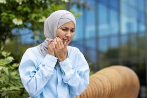 retrato do mulher vestindo hijab e luz azul camisa enquanto tocante bochecha com ambos mãos em fundo do vítreo construção exterior. apertando os olhos fêmea experimentando dentes sensibilidade sintoma. foto