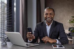 retrato do alegre maduro africano americano patrão dentro o negócio terno, Senior homem de negocios sorridente e olhando às Câmera com telefone dentro mãos, homem trabalhando dentro escritório com computador portátil, usando aplicativo. foto