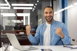 retrato do uma feliz e satisfeito jovem muçulmano homem sentado dentro a escritório às a mesa com uma computador portátil e alegria dentro sucesso, olhando às a Câmera e mostrando uma vitória gesto com dois mãos. foto