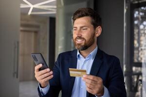 fechar-se foto do uma jovem homem de negocios em pé dentro a escritório Centro dentro uma terno, segurando uma crédito cartão e usando uma Móvel telefone.
