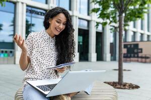 sorridente mulher trabalhando em dela computador portátil lado de fora a escritório prédio, acionado dentro a animado conversação em uma ensolarado dia. foto