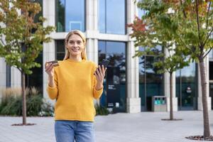 uma alegre Loiras mulher dentro uma amarelo suéter detém uma crédito cartão e uma Smartphone lado de fora uma moderno construção com árvores dentro a fundo. foto