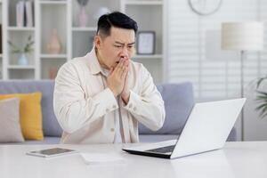 chocado ásia homem sentado às mesa às casa e olhando preocupado às computador portátil. detém mãos junto, mantido mau notícias, financeiro problemas, médico resultados. foto