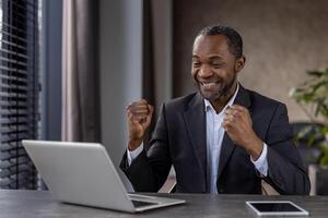 em êxtase africano americano homem de negocios punho bombeamento dentro triunfo às dele local de trabalho com uma computador portátil e tábua. foto