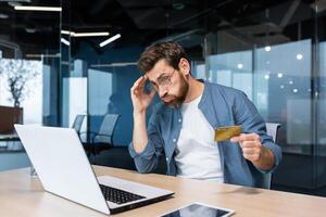 enganado e desapontado homem de negocios trabalhando dentro escritório, homem segurando banco crédito cartão e tentando para faço uma compra dentro conectados loja, conectados fraude e decepção. foto