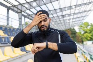 uma cansado masculino atleta lenços suor a partir de dele testa enquanto olhando às dele relógio inteligente, ilustrando exaustão e pessoal ginástica monitorando. foto