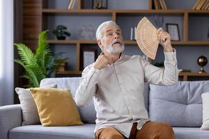 idosos caucasiano homem sentado em uma cinzento sofá, usando uma portátil ventilador para legal fora durante uma caloroso dia dentro de casa, olhando desconfortável e suado dentro uma bem decorado vivo sala. foto