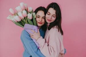 mãe abraços filha contra uma Rosa pano de fundo. mulher dentro azul suéter segurando tulipas, vestido dentro pastel Rosa foto