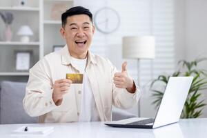 alegre ásia homem fazer gostar placa com mão enquanto sentado às doméstico escritório com computador portátil e plástico banco cartão. emocional controlo remoto programador recebendo financeiro bônus para produtivo por mês trabalhar. foto