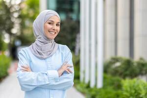 retrato do uma feliz e confiante muçulmano mulher vestindo uma hijab, sorridente calorosamente com braços cruzado dentro a urbano parque contexto. foto
