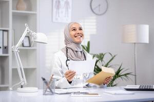 sorridente fêmea muçulmano médico dentro uma hijab e branco casaco segurando papéis dentro uma moderno clínica escritório, exibindo profissional assistência médica. foto